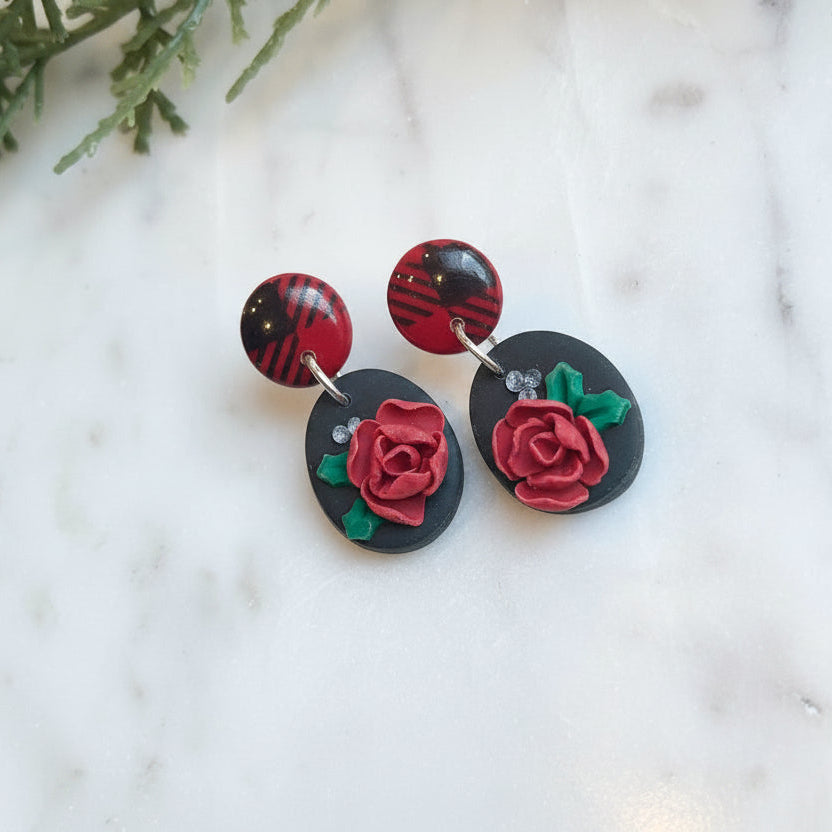 Pair of earrings with red rose design on a marble surface with greenery.